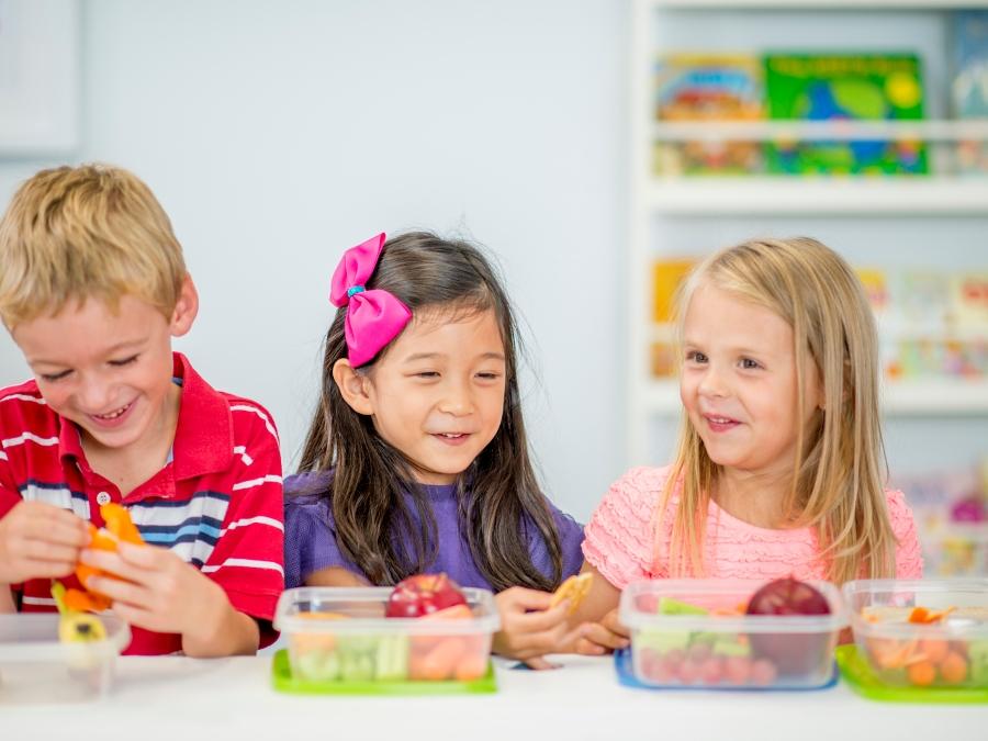 A multi ehthnic group of elementary age children are sitting together at a table eating their healthy snack full of apples, carrots, and celery.